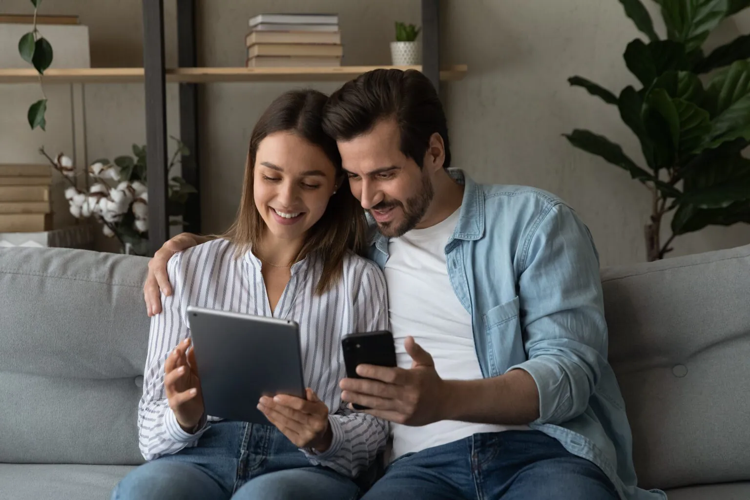 Ein lächelndes Paar sitzt auf einem Sofa, sieht gemeinsam auf ein Tablet und hält ein Smartphone.